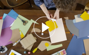 This image shows a preview of the 'What's the biodiversity challenge' video and includes a still shot of students doing craft activities on a table. There is a white play button in the middle ready to be clicked to launch the video.