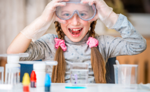 a young girl around 8 years old with platted pig-tails in her hair and pink scrunchies. She is wearing a pair of science goggles and is holding the sides of the goggles in protective gloves. She has a huge smile and looks excited. In the foreground, but not in focus, is science equipment like plastic beakers and solutions in bottles.